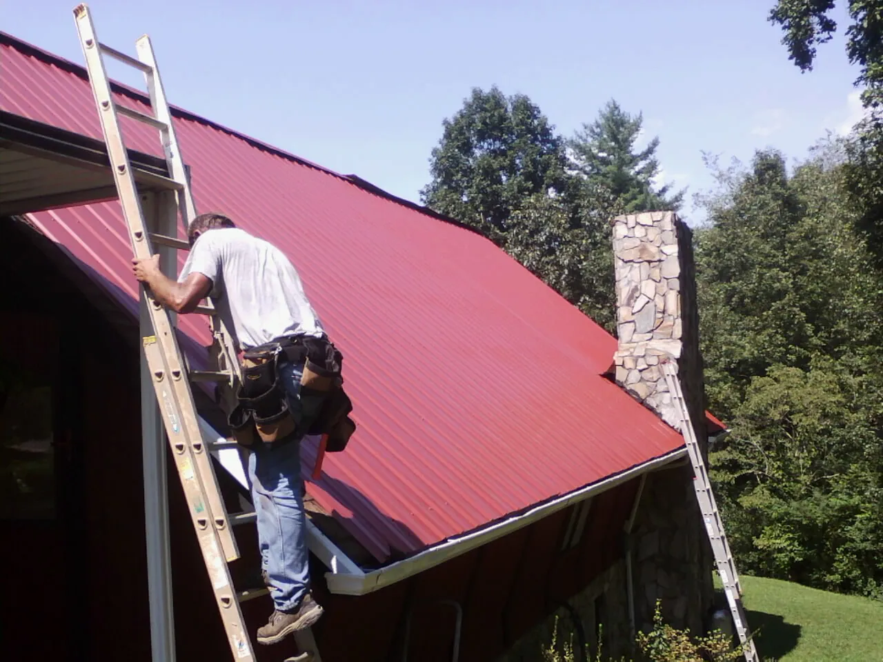 Roofer on a ladder beside a red metal roof and stone chimney — Soffit & Fascia Repair by Roofing Brothers in Pullman